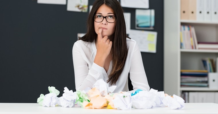 Young woman with writers block sitting in an office with a desk littered with crumpled paper as she sits looking thoughtfully into the air with her finger to her chin seeking new ideas